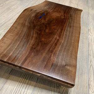 May include: A large, dark brown wooden slab with a live edge and a natural, organic shape. The wood has a rich, dark brown colour and a smooth, polished finish. There are two small blue resin inlays in the wood.