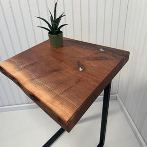 May include: A square side table featuring a natural live edge wood top with visible grain and small resin-filled knots, supported by a black metal C-shaped frame. A small green potted plant sits on the surface of the wooden tabletop.