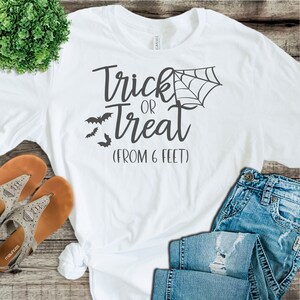 May include: White t-shirt with the text "Trick or Treat (From 6 Feet)" in black. The shirt also features a black spider web and bats.