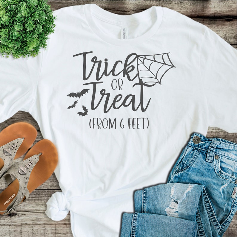 May include: White t-shirt with the text "Trick or Treat (From 6 Feet)" in black. The shirt also features a black spider web and bats.