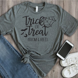 May include: A heather grey t-shirt with the text "Trick or Treat (From 6 Feet)" in black. The shirt features a spiderweb and bats.
