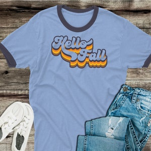 May include: A light blue t-shirt with a black ringer neck. The shirt features a retro-style graphic that says "Hello Fall" in orange, yellow, and blue.