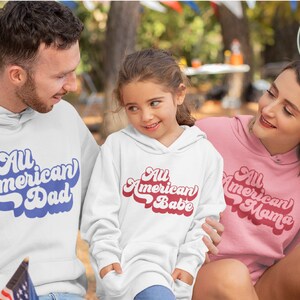May include: A family wearing white and pink hoodies with the text "All American Dad", "All American Babe", and "All American Mama" in a retro font. The family is sitting in a park setting.