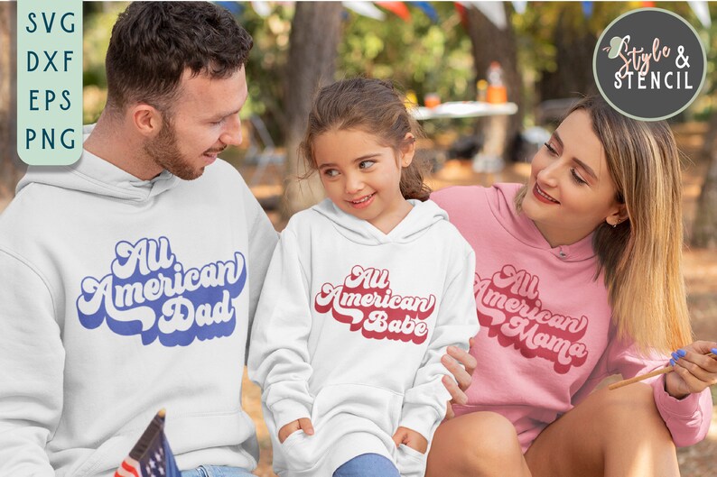 May include: A family wearing white and pink hoodies with the text "All American Dad", "All American Babe", and "All American Mama" in a retro font. The family is sitting in a park setting.