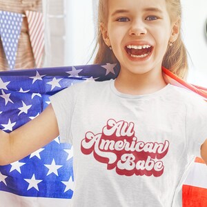 May include: A white t-shirt with red text that says "All American Babe" in a retro font. A young person is wearing the shirt and holding an American flag.
