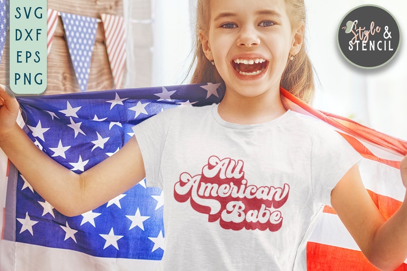 May include: A white t-shirt with red text that says "All American Babe" in a retro font. A young person is wearing the shirt and holding an American flag.