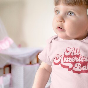 May include: A baby wearing a pink onesie with the text "All American Babe" in a retro font. The baby is looking to the right of the frame.