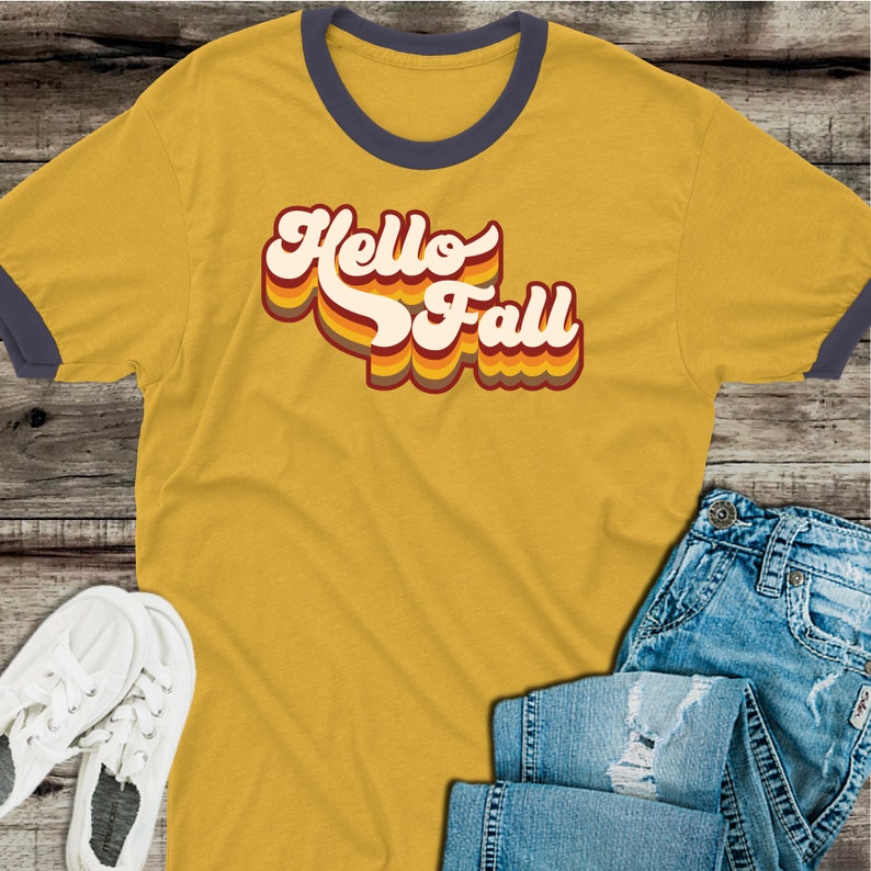 May include: A yellow t-shirt with a blue ringer neck. The shirt features a retro-style graphic that says "Hello Fall" in red, orange, and yellow.