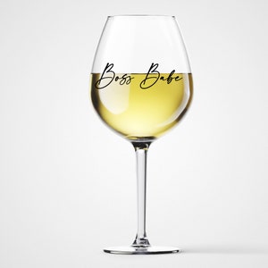 May include: A clear glass wine glass with a white wine inside. The glass has the text "Boss Babe" written on it.