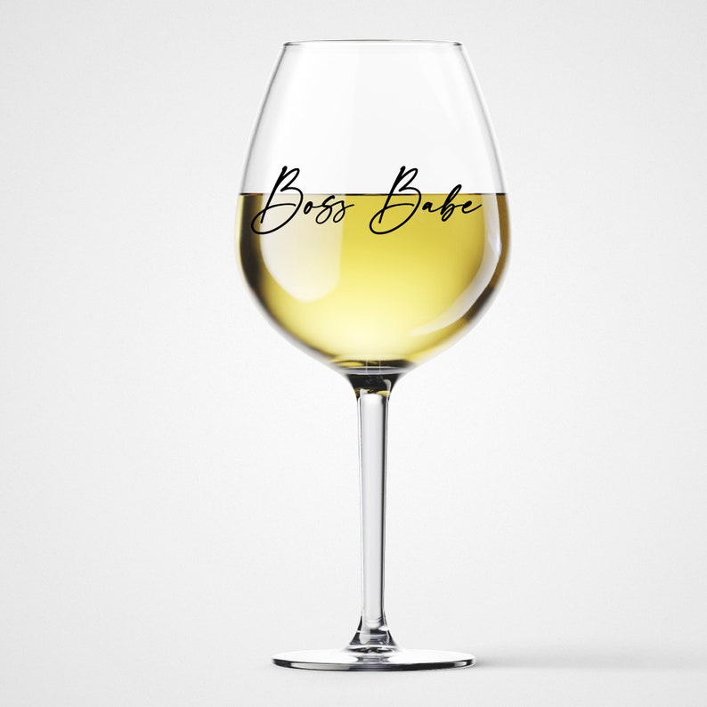 May include: A clear glass wine glass with a white wine inside. The glass has the text "Boss Babe" written on it.