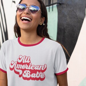 May include: A white t-shirt with a red ringer neckline and a red retro-style graphic that says "All American Babe".