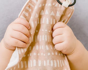 Peut inclure: Une sucette pour bébé attachée à un lange en mousseline à motifs. Le tissu présente un motif répété de formes blanches sur un fond beige. La sucette est transparente avec un anneau noir. Le tissu est tenu par un bébé.