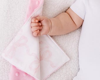Puede incluir: Una mantita de seguridad para bebé rosa y blanca con un chupete adjunto. La mantita presenta un lado rosa suave y texturizado con puntos en relieve y un lado blanco estampado con lazos rosas. El chupete es blanco con una tetina transparente.