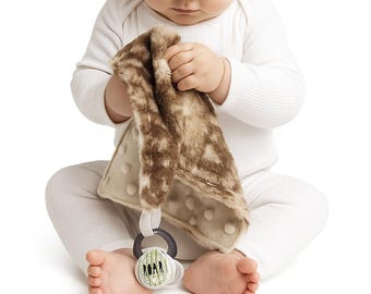 Peut inclure: Un bébé tient une couverture de sécurité en peluche marron et beige avec une surface texturée. La couverture est attachée à un hochet noir et blanc avec le mot "ROAR". Le bébé porte un body blanc. Un accessoire de confort pour bébé.