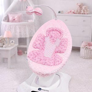 May include: A 4moms baby swing featuring a pink seat with a textured dot pattern and a Barbie-themed print. The seat includes a white harness, a curved overhead mobile with two pink plush balls, and a white base with a digital control panel.