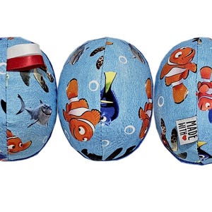 May include: Three blue, egg-shaped fabric ornaments with a cartoon fish theme. Each ornament features different characters, including orange clownfish, a blue tang, and a shark, on a light blue background. One ornament has a red and white ribbon.