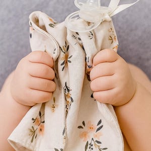 May include: A beige baby security blanket with a floral pattern in peach and green, held by a baby. A clear pacifier with a white strap is attached to the blanket. The blanket is soft and appears to be made of muslin.