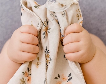 Peut inclure: Une couverture de sécurité pour bébé beige avec un motif floral pêche et vert, tenue par un bébé. Une sucette transparente avec une sangle blanche est attachée à la couverture. La couverture est douce et semble être en mousseline.