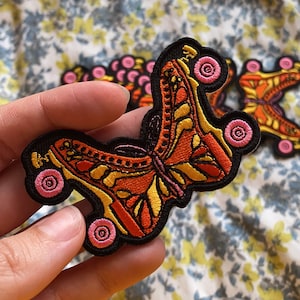 Op de afbeelding: Een close-up van een kleurrijk geborduurd patch met een vlinder met rolstoeltjes. De vlinder is oranje, geel en zwart met roze wielen. De patch is perfect om een unieke touch toe te voegen aan kleding, tassen of andere accessoires.