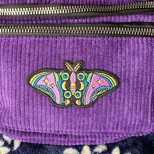 Könnte beinhalten: Lilafarbene Cord-Bauchtasche mit schwarzem Reißverschluss und einem bunten gestickten Motten-Patch.