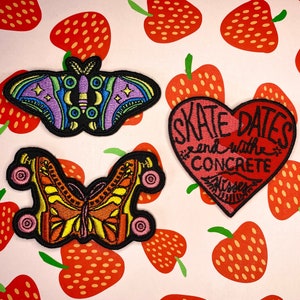 May include: Three embroidered iron-on patches. One is a pink heart with the text "Skate Dates end with Concrete". Another is a purple and blue butterfly with a yellow center. The last is a yellow and orange butterfly with a black outline and pink wheels.