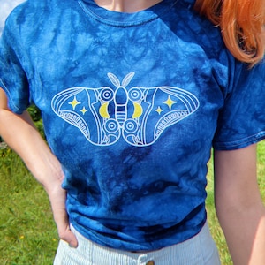 May include: A blue tie-dye t-shirt featuring a white moth design with yellow accents. The moth has detailed wings with stars and crescent moons. The shirt is a crew neck and has short sleeves. The background is a blue sky.