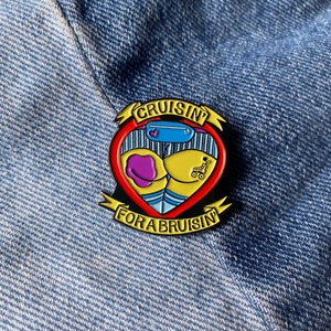 May include: A red heart-shaped enamel pin with a blue and white striped design, a purple heart, a yellow flower, and a small red wagon. The pin has a black border and the text "Cruisin' for a Bruisin'" in gold lettering.