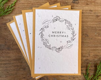 May include: A set of five white seed paper Christmas cards with a gold envelope. The cards feature a black and white illustration of a wreath with the text "Merry Christmas".