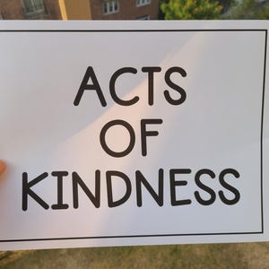 Puede incluir: Un letrero rectangular blanco con un borde negro muestra las palabras "ACTS OF KINDNESS" en negrita, con una fuente sans-serif. El letrero est&aacute; sostenido por una mano, con un fondo borroso al aire libre.
