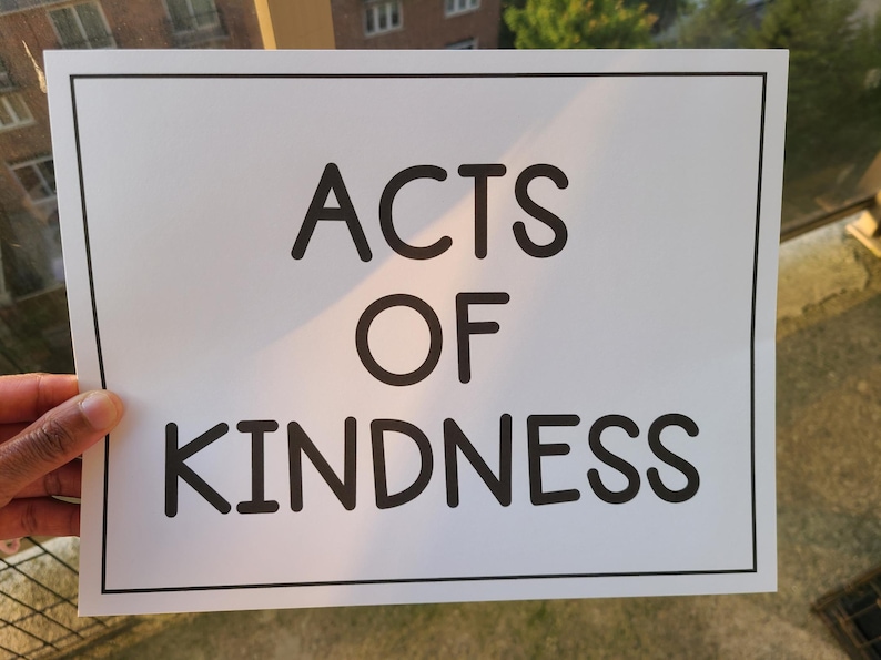 Puede incluir: Un letrero rectangular blanco con un borde negro muestra las palabras "ACTS OF KINDNESS" en negrita, con una fuente sans-serif. El letrero est&aacute; sostenido por una mano, con un fondo borroso al aire libre.