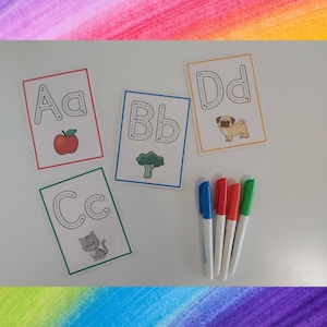 May include: Four white cards with black outlines of the letters A, B, C, and D. Each card has a picture of an apple, broccoli, cat, and pug. Four colorful dry-erase markers are on a white surface.