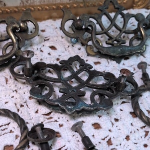 Peut inclure: Un ensemble de poignées de tiroir et de quincaillerie en laiton antique, doté de plaques arrière ajourées ornées de volutes complexes et de vis de montage assorties. Les surfaces métalliques présentent une patine vieillie aux tons sombres et verdâtres.