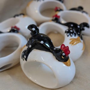 Könnte beinhalten: Eine Sammlung von Serviettenringen aus Keramik. Jeder Ring hat eine weiße Basis mit einem schwarz-weißen Hühnerdesign. Die Hühner haben rote Kämme und gelbe Akzente. Die Serviettenringe sind auf einer neutralfarbenen Oberfläche angeordnet.