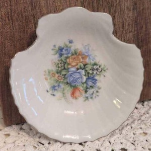 Può includere: Un piatto in porcellana bianca a forma di conchiglia con bordo smerlato e finitura color argento. Il centro del piatto è decorato con un'illustrazione di un bouquet floreale con fiori e foglie blu, arancioni e verdi su sfondo bianco.