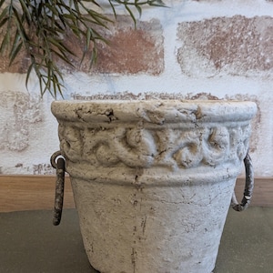 Puede incluir: Pequeña maceta blanquecina, con aspecto envejecido, con un patrón decorativo en relieve alrededor del borde. Tiene dos asas metálicas y un acabado desgastado. Adecuada para plantas pequeñas, con un estilo rústico.