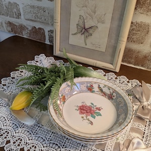May include: A stack of four decorative ceramic bowls with a floral and bird design. The bowls have a white interior with a red flower and a blue and gold rim. The bowls are on a white lace tablecloth.
