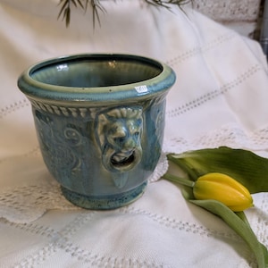 May include: A blue ceramic planter with a lion's head handle. The planter has a round shape with a decorative rim and base. A yellow tulip rests on the white lace tablecloth.