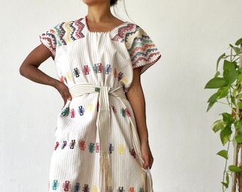 May include: A white cotton dress with colorful embroidered details and a striped pattern. The dress has short sleeves and a tie belt. The model is wearing the dress with brown sandals.