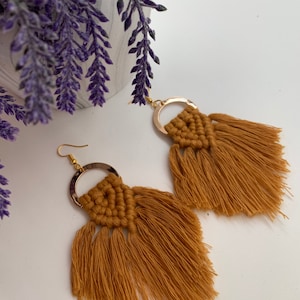May include: A pair of mustard yellow macrame earrings featuring a gold-toned metal hoop at the top. The cotton cord is knotted in a triangular pattern, extending into long, loose fringe tassels. Each earring includes a gold-toned metal hook.