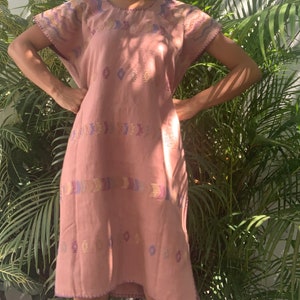 Pode incluir: Um vestido rosa bordado com decote redondo e mangas curtas. O vestido tem um padrão geométrico em tons de roxo, azul, amarelo e dourado.
