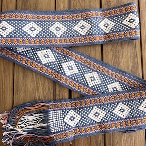 May include: A woven belt with a blue background and a repeating pattern of white diamonds and orange and pink accents. The belt has a fringed end with blue, orange, and white threads. The belt is displayed on a wooden surface.