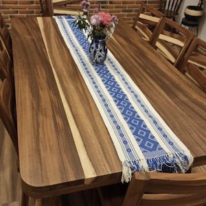 Peut inclure: Une table à manger en bois avec un chemin de table tissé bleu et blanc. La table a une finition en bois naturel et est entourée de chaises en bois.