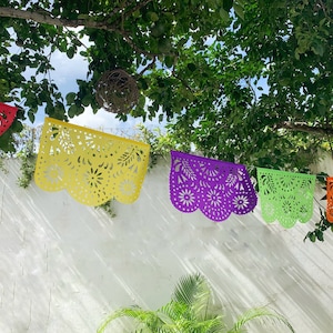 May include: Five colorful paper banners with intricate cut-out designs hang from a tree branch against a white wall. The banners are red, yellow, purple, green, and orange.