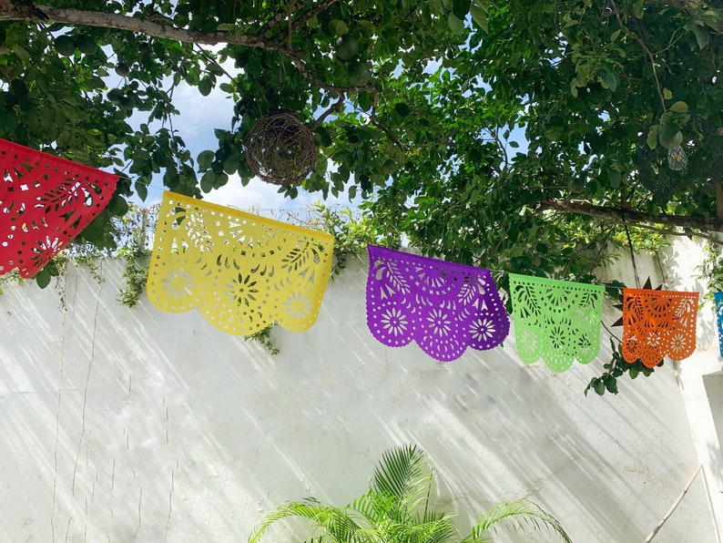 May include: Five colorful paper banners with intricate cut-out designs hang from a tree branch against a white wall. The banners are red, yellow, purple, green, and orange.
