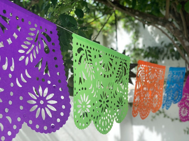 May include: A string of colorful paper banners with intricate cut-out designs. The banners are purple, green, orange, and blue. They are hanging from a string and are strung together in a row.