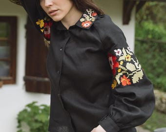 May include: Black button-down shirt with floral embroidery on the collar and sleeves. The sleeves are long and gathered at the cuffs. The shirt is paired with light wash denim jeans. The embroidery features red, yellow, and green floral patterns.