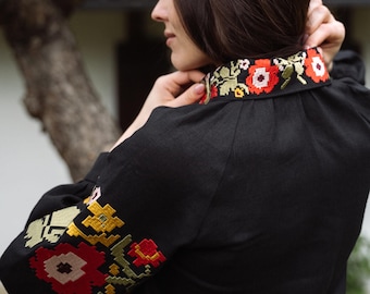 May include: Black linen blouse with colorful floral embroidery on the collar and sleeves. The embroidery features red, yellow, and green flowers. The blouse has long sleeves and a high neckline.