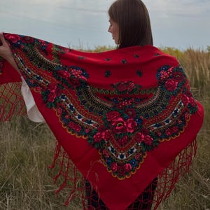 Peut inclure: Un grand châle carré rouge orné d'un motif floral vibrant composé de roses dans des tons de rose, rouge et blanc, agrémenté de feuilles vertes et de bordures décoratives noires et dorées. Les bords sont finis par de longues franges rouges nouées.