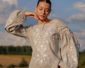 May include: A gray linen blouse with white embroidered floral designs. The blouse has long sleeves with gathered cuffs and a round neckline.