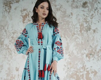 May include: A light blue embroidered dress with red and blue floral patterns. The dress has long sleeves, a round neckline, and a tie belt. The dress is made of linen and has a flowing skirt.